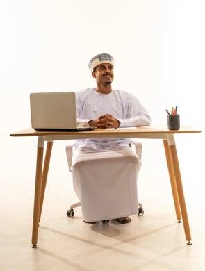 دمج التقنيات الحديثة بالمجال الإداري والمكتبي، العمل في المكاتب والشركات العمانية، بورتريه لرجل عربي خليجي عماني يرتدي الدشداشه والعمامه يجلس خلف طاولة المكتب امامه جهاز الحاسوب المحمول و يقوم بالنظر الى شيء ما بايماءات السعادة و السرور، خلفية بيضاء