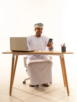 إدخال التقنيات المتطورة في روتين الحياة، الدردشة والتواصل مع الأهل والأصدقاء وسماع أخبارهم، بورتريه لرجل عربي خليجي عماني يرتدي الدشداشة و العمامة يجلس خلف طاولة خشبية وامامه حاسوب محمول يقوم بتصفح الجوال,ايماءات السعادة و السرور, خلفية بيضاء