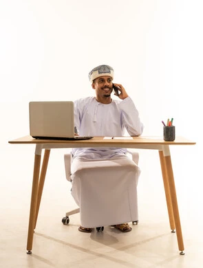 اجراء مكالمة هاتفيه عبر الجوال، الدردشة مع الأقارب والأصدقاء وتبادل الأخبار، دمج التكنولوجيا المتطورة بالحياة اليومية، بورتريه لرجل عربي خليجي عماني يرتدي الدشداشة و العمامة يجلس خلف طاولة خشبية وامامه حاسوب محمول يمسك بيده الهاتف المحمول, استخدام جهاز تقني حديث, خلفية بيضاء