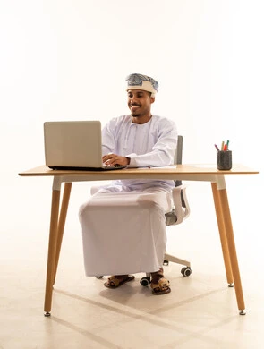متابعة وتنظيم الأعمال الإدارية ، استخدام الأجهزة التكنولوجية الحديثة والمتطورة، بورتريه لرجل عربي خليجي عماني يرتدي الدشداشة و العمامة يجلس خلف طاولة خشبية وامامه حاسوب محمول يقوم بالكتابة والنقر على لوحة المفاتيح , ايماءات السعادة و السرور، خلفية بيضاء