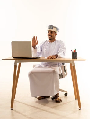 الاستفادة من التقنيات الحديثة في القطاع المكتبي والإداري، اجراء مكالمة فيديو عمل عبر الابتوب, إيماءات يدين تدل على الترحيب، بورتريه لرجل عربي خليجي عماني يرتدي الدشداشه و العمامة  يجلس خلف طاولة المكتب يدير العمل عن بعد، خلفية بيضاء