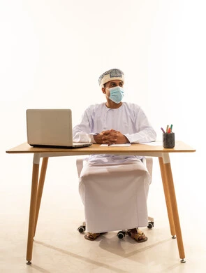 استخدام الأجهزة الحديثة والتقنيات المتطورة، مفهوم ادارة الاعمال عن بعد، مهن ووظائف مكتبيه عمانية، بورتريه لشاب عربي خليجي عماني يرتدي الدشداشة والعمامة و الكمامة يجلس خلف طاولة خشبية وامامه حاسوب محمول ينظر الى شيء ما، خلفية بيضاء