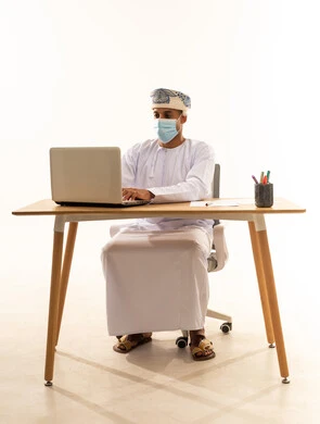 إدارة شؤون الأعمال المكتبية وتنظيمها، استخدام الأجهزة التكنولوجية الحديثة والمتطورة، بورتريه لرجل عربي خليجي عماني يرتدي الدشداشة و العمامة و الكمامة يجلس خلف طاولة خشبية وامامه حاسوب محمول يقوم بالكتابة والنقر على لوحة المفاتيح , خلفية بيضاء