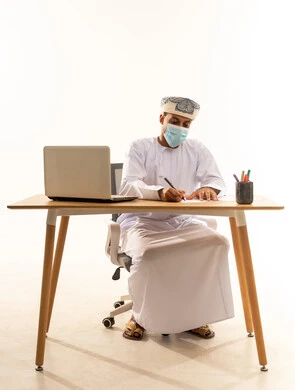تسجيل الملاحظات والمعلومات المهمة، استخدام التقنيات الحديثة ، بورتريه لرجل عربي خليجي عماني يرتدي الدشداشة و العمامة و الكمامة يجلس خلف طاولة خشبية وامامه حاسوب محمول يقوم بكتابة شي ما , خلفية بيضاء