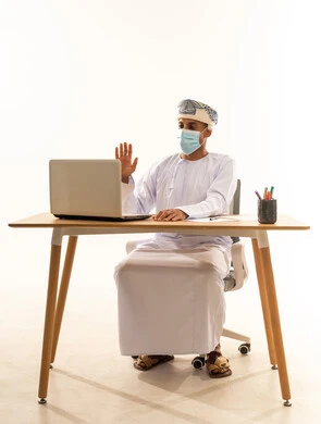 إجراء مكالمة فيديو عبر جهاز الحاسوب المحمول، استخدام التقنيات الحديثة في التواصل مع العملاء، بورتريه لرجل عربي خليجي عماني يرتدي الدشداشة و العمامة و الكمامة يجلس خلف طاولة خشبية وامامه حاسوب محمول يرفع يديه بايماءات القاء التحية والسلام, خلفية بيضاء