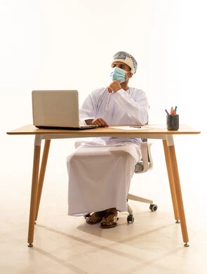 التأمل والتخطيط للمستقبل، إيماءات محاولة تذكر أمر ما، ، بورتريه لشاب عربي خليجي عماني يرتدي الدشداشة و العمامة والكمامة يجلس خلف طاولة خشبية وامامه حاسوب محمول يضع يده على ذقنه ينظر إلى الأعلى بتعابير التفكير، التركيز والتمعن لإيجاد حل لمشكلة معينة، خلفية بيضاء