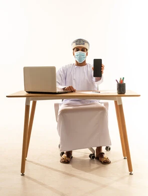 النظر الى الكاميرا بتعابير السرور، استخدام التكنولوجيا و التقنيات الحديثة المتطورة، بورتريه لشاب عربي خليجي عماني يرتدي الدشداشة و العمامة والكمامة يجلس خلف طاولة خشبية وامامه حاسوب محمول يقوم بعرض شاشة سوداء فارغة عبر الهاتف المحمول، خلفية بيضاء