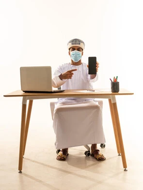 الاشارة الى شاشة سوداء فارغه، استخدام التكنولوجيا و التقنيات الحديثة المتطورة، بورتريه لشاب عربي خليجي عماني يرتدي الدشداشة و العمامة والكمامة يجلس خلف طاولة خشبية وامامه حاسوب محمول يمسك بيده الهاتف المحمول، خلفية بيضاء