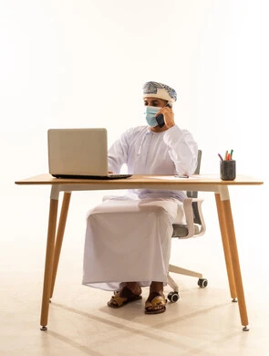 Exchanging news with family and friends, using modern devices and advanced technologies, the concept of remote business management, Omani office professions and jobs, a portrait of an Arab Gulf Omani man wearing a dishdasha and turban and a coronavirus protection mask making a phone call via a mobile phone, white background.