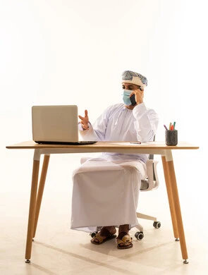 استخدام الأجهزة الحديثة والتقنيات المتطورة، مفهوم ادارة الاعمال عن بعد، مهن ووظائف مكتبيه عمانية, بورتريه لرجل عربي خليجي عماني يرتدي الدشداشة والعمامة و قناع حماية كورونا يقوم باجراء مكالمة هاتفية عبر الهاتف المحمول, خلفية بيضاء