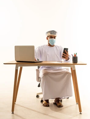 الدردشة مع الأقارب والأصدقاء وتبادل الأخبار، دمج التكنولوجيا المتطورة بالحياة اليومية، بورتريه لرجل عربي خليجي عماني يرتدي الدشداشة و العمامة و قناع حماية كورونا يجلس خلف طاولة خشبية وامامه حاسوب محمول يقوم بتصفح الهاتف المحمول, استخدام جهاز تقني حديث, خلفية بيضاء