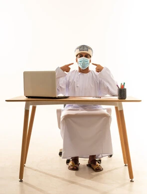 استخدام جهاز تقني حديث، دمج التكنولوجيا المتطورة بالحياة اليومية، بورتريه لرجل عربي خليجي عماني يرتدي الدشداشة و العمامة والكمامة يجلس خلف طاولة خشبية وامامه حاسوب محمول يقوم بالاشارة الى وجهه,النظر الى الكاميرا بايماءات السرور، خلفية بيضاء