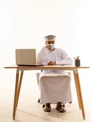 التمعن والتركيز لإيجاد الحل لمشكلة ما, الشعور بالضيق والملل، حل المشكلات واتخاذ القرارات، بورتريه لشاب عربي خليجي عماني يرتدي الدشداشة و العمامة و الكمامة يجلس خلف طاولة خشبية وامامه حاسوب محمول يضع يده على ذقنة بإيماءات التفكير والحيرة والانزعاج، خلفية بيضاء