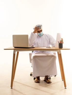 التركيز وشرود الذهن والتمعن، بورتريه لشاب عربي خليجي عماني يرتدي الدشداشة و العمامة و الكمامة يجلس خلف طاولة خشبية وامامه حاسوب محمول يمسك بيده اوراق و يقوم بالتفكير فى شيء ما، خلفية بيضاء