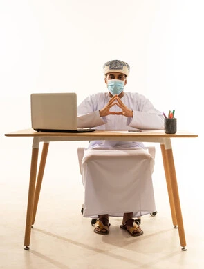 النظر الى الكاميرا يايماءات الثقة بالنفس، تعابير السعادة والسرور، مفهوم الذكاء والمعرفة، بورتريه لشاب عربي خليجي عماني يرتدي الدشداشة و العمامة والكمامة يجلس خلف طاولة خشبية وامامه حاسوب محمول يرفع يديه بحركة تلامس الأصابع، خلفية بيضاء