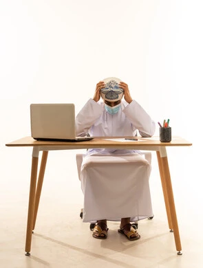 Fear, anxiety, and tension, a portrait of a young Arab Gulf Omani man wearing a dishdasha, turban, and mask sitting behind a wooden table with a laptop in front of him. He sits in front of the laptop with his hands on his head, showing gestures of discomfort and nervousness, conveying a sense of disappointment, with a white background.