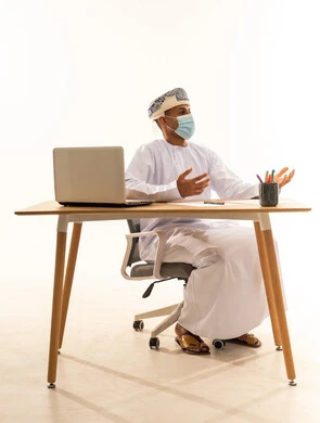 مفهوم الجد والاجتهاد في إنجاز المهام المطلوبة، اجراء مكالمة فيديو عبر الابتوب، بورتريه لشاب عربي خليجي عماني يرتدي الدشداشة و العمامة والكمامة يجلس خلف طاولة خشبية وامامه حاسوب محمول يقوم بشرح شيء ما، خلفية بيضاء