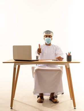 الاعجاب والرضا لتحقيق أمر معين، ادارة الاعمال عن بعد، مهن ووظائف مكتبيه عمانية، بورتريه لشاب عربي خليجي عماني يرتدي الدشداشة والعمامة و قناع حماية كورونا يجلس خلف طاولة خشبية وامامه حاسوب محمول يرفع اصبع الإبهام إلى الاعلى بعلامة الجودة والامتياز، خلفية بيضاء