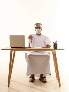 مفهوم الرفض وعدم القبول، ادارة الاعمال عن بعد، مهن ووظائف مكتبيه عمانية، بورتريه لشاب عربي خليجي عماني يرتدي الدشداشة والعمامة و قناع حماية كورونا يجلس خلف طاولة خشبية وامامه حاسوب محمول يشير بإصبع الابهام للاسفل بإيماءات عدم الاعجاب والتقييم السلبي، خلفية بيضاء