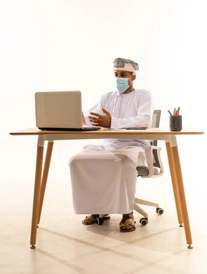 إجراء مكالمة فيديو عبرالابتوب، استخدام الأجهزة الحديثة والتقنيات المتطورة، مفهوم ادارة الاعمال عن بعد، مهن ووظائف مكتبيه عمانية، بورتريه لشاب عربي خليجي عماني يرتدي الدشداشة والعمامة و قناع حماية كورونا يجلس خلف طاولة خشبية وامامه حاسوب محمول، خلفية بيضاء