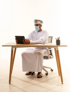 الاستفادة من التقنيات الحديثة في القطاع المكتبي والإداري، استخدام الجهاز اللوحي في الإشراف على سير الأعمال، مهن ووظائف في المكاتب والشركات العمانية، بورتريه لرجل عربي خليجي عماني يرتدي الدشداشه و العمامة والكمامة يجلس خلف طاولة المكتب يدير العمل عن بعد، خلفية بيضاء