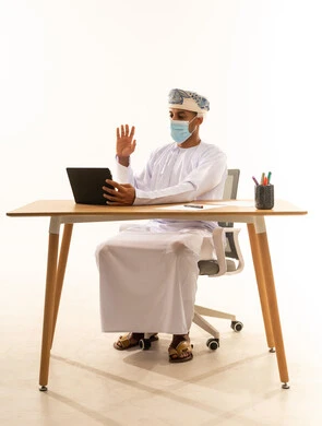 التحدث مع الأهل و الأصدقاء عبر الجهاز اللوحي, إيماءات يدين تدل على الترحيب، الاستفادة من التقنيات الحديثة في القطاع المكتبي والإداري، بورتريه لرجل عربي خليجي عماني يرتدي الدشداشه و العمامة و الكمامة يجلس خلف طاولة المكتب يدير العمل عن بعد، خلفية بيضاء