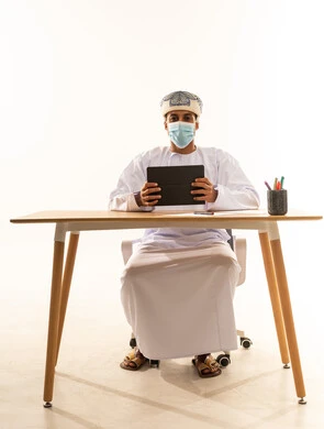 The use of a tablet in supervising the workflow, professions and jobs in Omani offices and companies, benefiting from modern technologies in the office and administrative sector, a portrait of an Arab Gulf Omani man wearing a turban, dishdasha, and mask sitting behind a desk managing work remotely, white background.