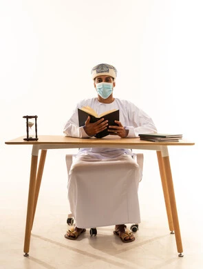 الاستمتاع بممارسة هواية المطالعة، مفهوم التثقيف والبحث العلمي، بورتريه لرجل عربي خليجي عماني يرتدي العمامة والدشداشة والكمامة يجلس على كرسي و طاولة يقوم بقراءة كتاب، خلفية بيضاء