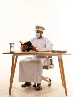 الأنشطة الترفيهية المفيدة، مفهوم التثقيف والبحث العلمي، هواية المطالعة والاستكشاف، بورتريه لرجل عربي خليجي عماني يرتدي العمامة والدشداشة والكمامة يجلس على كرسي و طاولة يقوم بقراءة كتاب، خلفية بيضاء