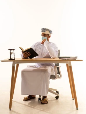 قضاء وقت الفراغ بالتعلم والثقافة، مفهوم التثقيف والبحث العلمي، هواية المطالعة والاستكشاف، بورتريه لرجل عربي خليجي عماني يرتدي العمامة والدشداشة والكمامة يجلس على كرسي و طاولة يقوم بقراءة كتاب، خلفية بيضاء