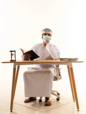 مفهوم التثقيف والبحث العلمي، هواية المطالعة والاستكشاف، قضاء وقت الفراغ بالتعلم والثقافة، بورتريه لرجل عربي خليجي عماني يرتدي العمامة والدشداشة والكمامةا يجلس على كرسي و طاولة يمسك كتاب ويضع يده على ذقنه ينظر إلى شيء ما بتعابير التفكير، خلفية بيضاء