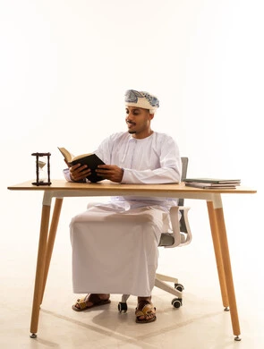 The hobby of reading and exploration, gestures of immersion and focus, the concept of education and scientific research, spending free time learning and cultural activities, a portrait of an Arab Gulf Omani man wearing a kumma and dishdasha sitting on a chair at a table reading a book, white background.