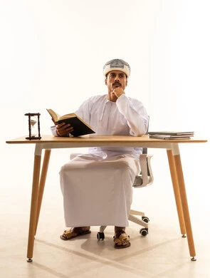 Gestures of thinking about something, the concept of education and scientific research, the hobby of reading and exploration, spending free time on learning and culture, a portrait of an Arab Gulf Omani man wearing a turban and a dishdasha sitting on a chair and table reading a book, white background.