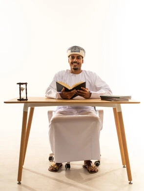 النظر الى الكاميرا بايماءات السعادة و السرور، مفهوم التثقيف والبحث العلمي، هواية المطالعة والاستكشاف، قضاء وقت الفراغ بالتعلم والثقافة، بورتريه لرجل عربي خليجي عماني مبتسم يرتدي العمامة والدشداشة يجلس على كرسي و طاولة يقوم بقراءة كتاب، خلفية بيضاء