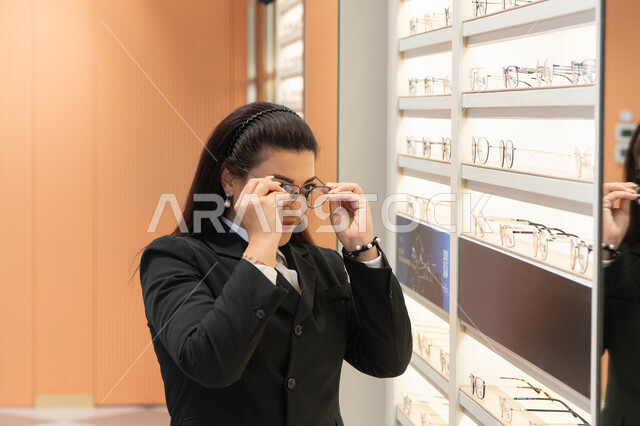 الأناقة والاهتمام بالمظهر الخارجي، مفهوم الرؤية الواضحة، ماركات عالمية فاخرة، صورة مقربة لسيدة عربية خليجية اماراتيه ترتدي الزي الرسمي تقوم باختيار نظارة ملائمة لملامح الوجه، امرأة سعوديه تقوم بمعاينة النظارات، الاعجاب بالنظارات الطبية الجديدة، ضعف ونقص النظر