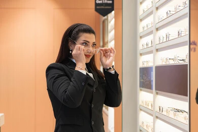 تصحيح وتحسين الرؤية، التسوق والشراء من المحلات الإماراتية، تعابير الثقة بالنفس، مفهوم ضعف النظر، اقتناء نظارات طبية جديدة، صورة مقربة لسيدة عربية خليجية سعودية ترتدي الزي الرسمي تقوم باختيار نظارة ملائمة لملامح الوجه، امرأة إماراتية تقوم بمعاينة النظارات