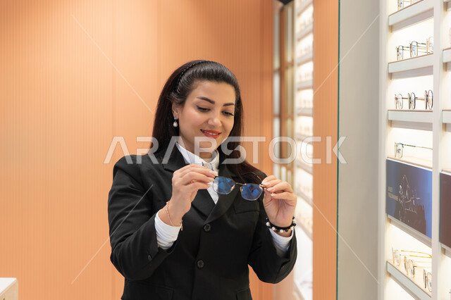 ماركات عالمية فاخرة، تحسين وتصحيح الرؤية، مفهوم ضعف النظر، صورة مقربة لسيدة عربية خليجية اماراتيه ترتدي الزي الرسمي تقوم باختيار نظارة ملائمة لملامح الوجه، امرأة سعوديه تقوم بمعاينة النظارات، الاعجاب بالنظارات الطبية الجديدة،