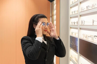 تصحيح وتحسين الرؤية، التسوق والشراء من المحلات الإماراتية، تعابير الثقة بالنفس، مفهوم ضعف النظر، اقتناء نظارات طبية جديدة، صورة مقربة لسيدة عربية خليجية سعودية ترتدي الزي الرسمي تقوم باختيار نظارة ملائمة لملامح الوجه، امرأة إماراتية تقوم بمعاينة النظارات