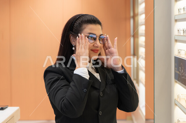 التسوق والشراء من المحلات الإماراتية، تعابير الثقة بالنفس، مفهوم ضعف النظر، اقتناء نظارات طبية جديدة، صورة مقربة لسيدة عربية خليجية سعودية ترتدي الزي الرسمي تقوم باختيار نظارة ملائمة لملامح الوجه، امرأة إماراتية تقوم بمعاينة النظارات