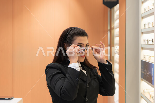 استخدام البصريات لتوضيح الرؤية، محاولة تعديل وتحسين البصر، شابة عربية خليجية سعودية ترتدي الزي الرسمي تقوم باختيار نظارة طبية ملائمة لملامح الوجه، صورة مقربة لامرأة اماراتية تقوم بشراء النظارات الخاصة بضعف وقصرالنظر، متعة التسوق في المتاجر الإماراتية