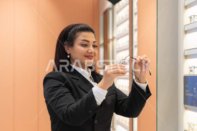 Using optics to clarify vision, trying to modify and improve eyesight, a young Saudi Arabian Gulf woman wearing a formal uniform choosing eyeglasses that fit her facial features, a close-up of an Emirati woman buying glasses for nearsightedness and myopia, the joy of shopping in Emirati stores