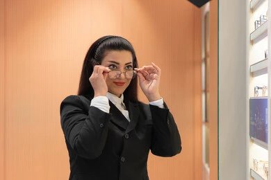 التسوق والشراء من المحلات الإماراتية، تعابير الثقة بالنفس، مفهوم ضعف النظر، اقتناء نظارات طبية جديدة، صورة مقربة لسيدة عربية خليجية سعودية ترتدي الزي الرسمي تقوم باختيار نظارة ملائمة لملامح الوجه، امرأة إماراتية تقوم بمعاينة النظارات