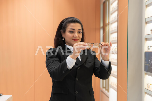 الاعجاب بالنظارات الطبية الجديدة، تصحيح وتحسين الرؤية، مفهوم ضعف النظر، صورة مقربة لسيدة عربية خليجية سعودية ترتدي الزي الرسمي تقوم باختيار نظارة ملائمة لملامح الوجه، امرأة إماراتية تقوم بمعاينة النظارات، متعة التسوق والشراء من المحلات الإماراتية