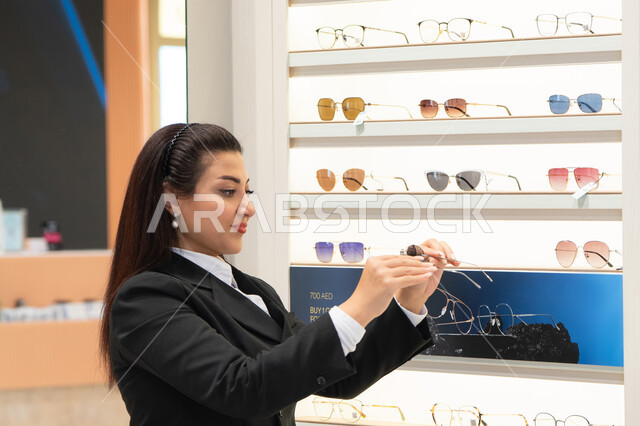 The latest stylish and modern women's accessories, the pleasure and enjoyment of shopping in the Emirates malls, keeping up with the latest fashion and beauty trends, a close-up of an Emirati Gulf Arab woman wearing formal attire choosing sunglasses that suit her facial features, a Saudi woman examining glasses