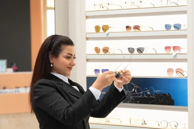 التركيز والتمعن لاختيار اللون المناسب، متابعة آخر صيحات الموضة، أحدث الاكسسوارات النسائية الأنيقة، التسوق في مولات الإمارات، صورة مقربة لسيدة عربية خليجية اماراتية ترتدي الزي الرسمي تقوم باختيار نظارة شمسية ملائمة لملامح الوجه، امرأة سعودية تقوم بمعاينة النظارات