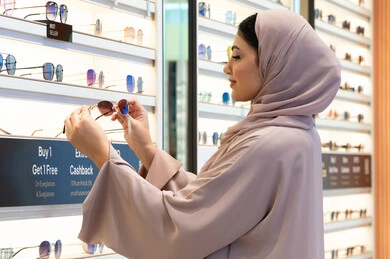 أحدث الاكسسوارات النسائية العصرية الأنيقة، متابعة آخر صيحات الموضة والجمال، متعة التسوق في مولات الإمارات، امرأة سعودية تقوم بمعاينة النظارات الملونة، صورة من الجانب لشابة عربية خليجية اماراتية محجبة ترتدي عباءة تقوم باختيار نظارة شمسية ملائمة لملامح الوجه