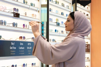 Emirati store selling the latest women's accessories, enjoy shopping in the UAE malls, keep up with the latest fashion and beauty trends, a close-up side view of a veiled Emirati Gulf Arab young woman wearing an abaya choosing sunglasses that suit her facial features, a Saudi woman examining the glasses