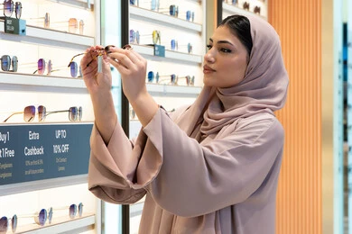 قضاء وقت ممتع بالتسوق في مولات الإمارات، مواكبة آخر صيحات الموضة والجمال، أحدث الاكسسوارات النسائية العصرية الأنيقة، صورة من الجانب لشابة عربية خليجية اماراتية محجبة ترتدي عباءة تقوم باختيار نظارة شمسية ملائمة لملامح الوجه،  امرأة سعودية تقوم بمعاينة النظارات