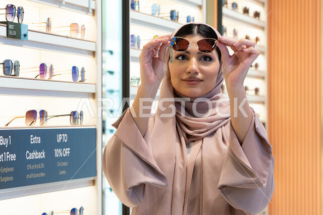 شراء اكسسوارات نسائية عصرية أنيقة، متابعة آخر صيحات الموضة والجمال، متعة التسوق في مولات الإمارات، امرأة سعودية تقوم بمعاينة النظارات الملونة، شابة عربية خليجية اماراتية محجبة ترتدي عباءة تقوم باختيار نظارة شمسية ملائمة لملامح الوجه