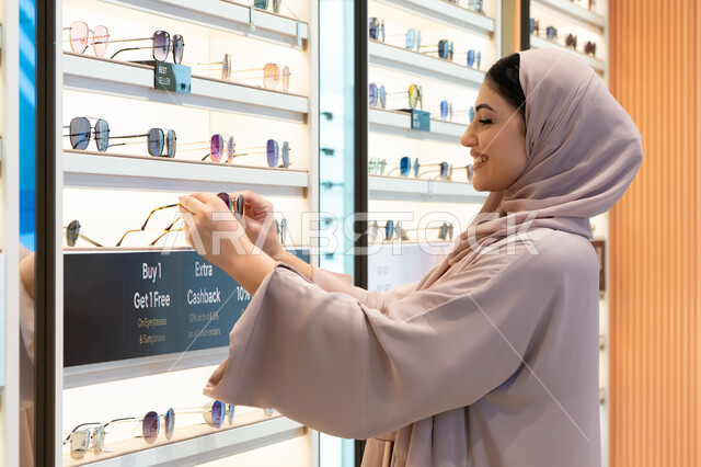 الاهتمام بالمظهر الخارجي، أحدث الاكسسوارات النسائية، المتعة بالتسوق في مولات الإمارات، مواكبة آخر صيحات الموضة والجمال، شابة عربية خليجية اماراتية محجبة ترتدي عباءة تقوم باختيار نظارة شمسية ملائمة لملامح الوجه،  امرأة سعودية تقوم بمعاينة النظارات